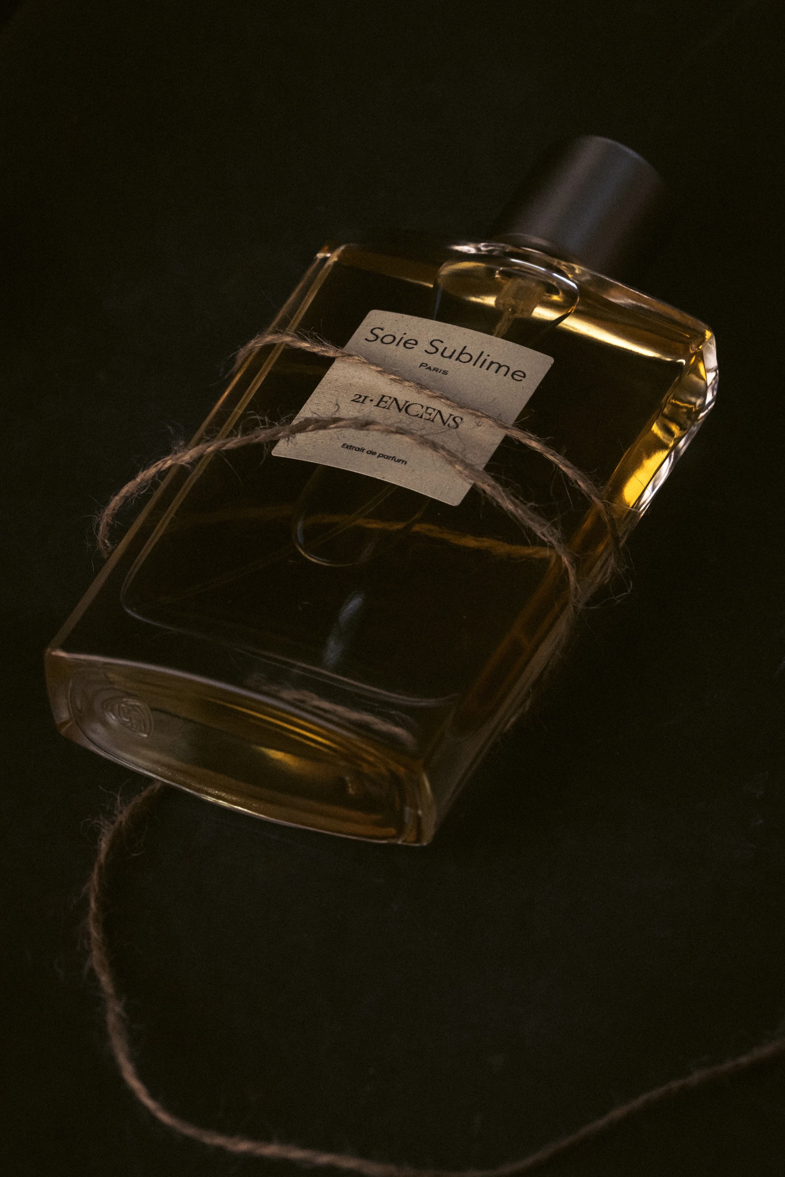 Flacon ENCENS couché sur un fond noir, enlacé d’une ficelle de jute dans un clair-obscur ambré